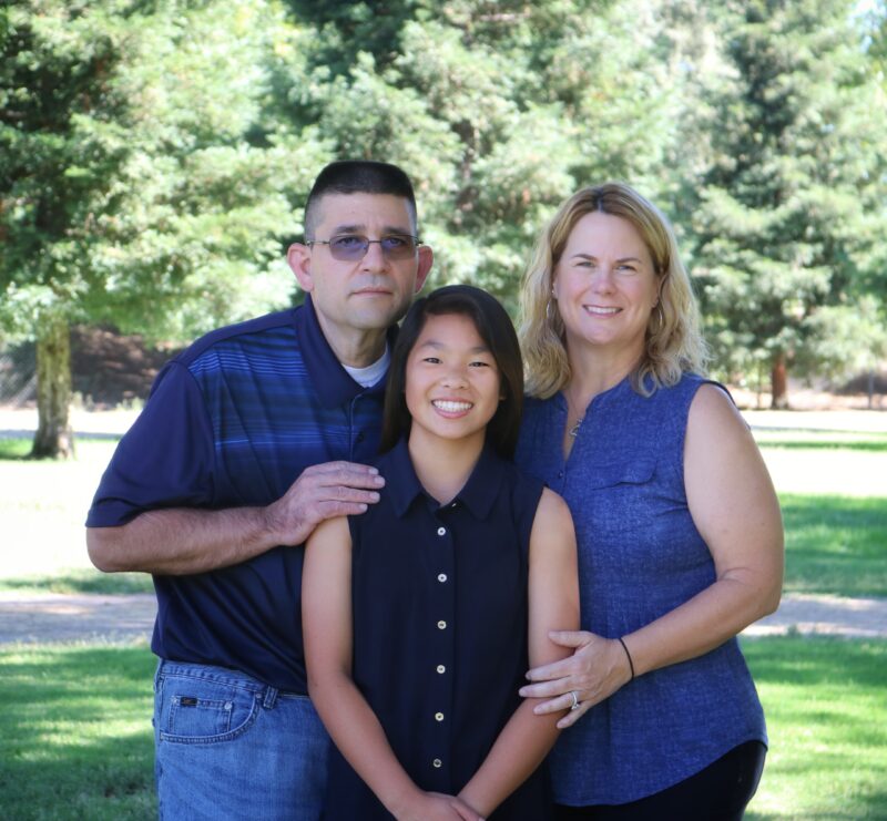 An image of Amy posing with her family outside with a brightly lit copse of trees in the background.